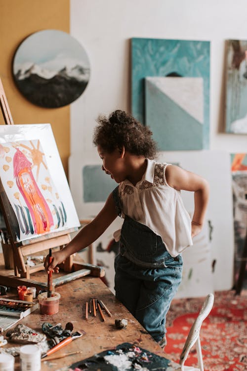 A Girl Holding a Paint Brush in a Container with Water Beside a Painting
