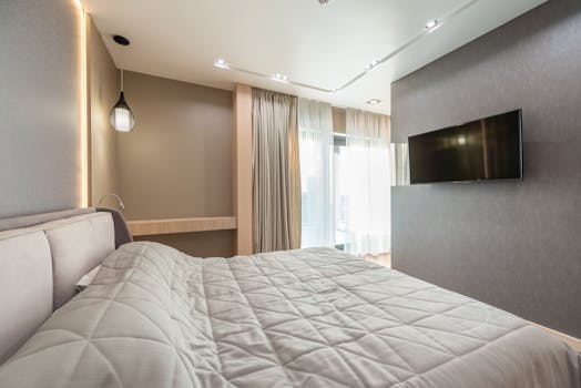Soft bed covered with quilted blanket placed in front of TV hanging on gray wall in modern bedroom