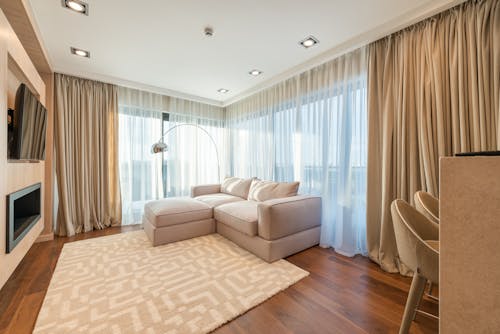 Comfortable sofa with cushions placed near windows covered with curtains in living room with kitchen set and soft carpet