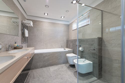 White bidet placed near bath against sinks bear liquid soap and mirror in stylish bathroom with glass wall in apartment