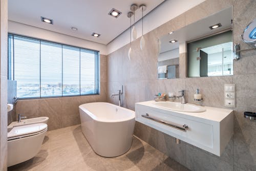 White bathtub and bidet placed near sink with hygienic supplies and mirror in stylish light bathroom with jalousie on window
