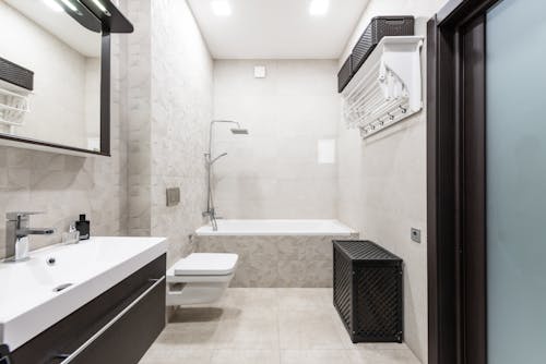 Bath with shower head placed near white toilet and sink near mirror in stylish light bathroom with door and black containers
