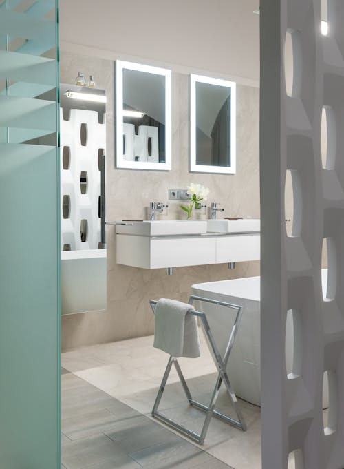 Interior of modern bathroom with white bath and sinks near illuminated mirrors against decorative partition and glass door in apartment