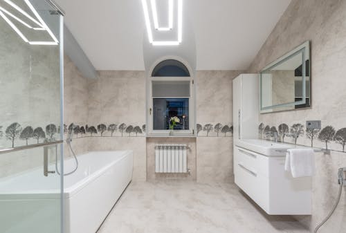 White bath and sink with mirror placed near glass shower cabin and window in spacious stylish bathroom with glowing illumination on ceiling