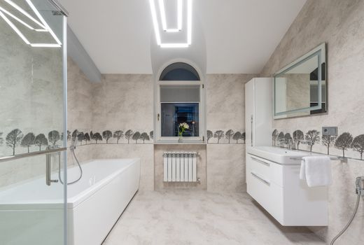 White bath and sink with mirror placed near glass shower cabin and window in spacious stylish bathroom with glowing illumination on ceiling