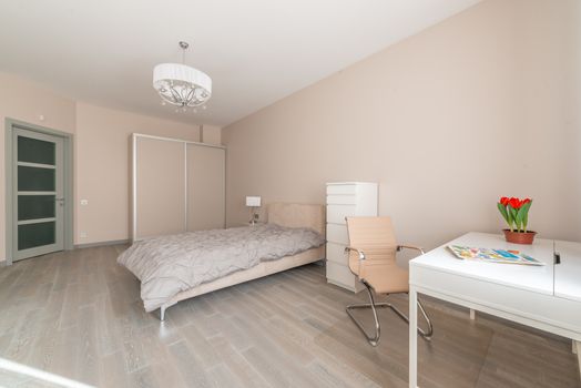 Comfortable bed placed between wardrobe and cupboard in spacious bedroom with door and chair at white table with flowerpot