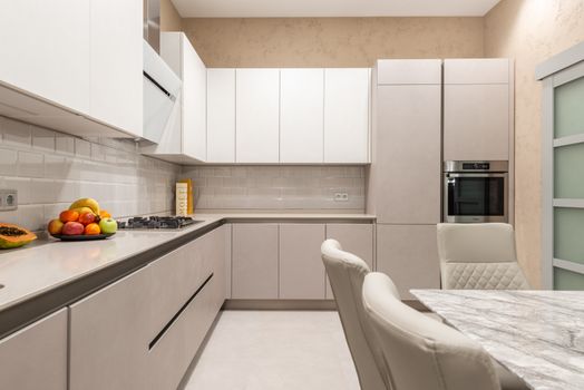 Table with chairs placed near cupboards and counter with stove and fruits in stylish kitchen with modern kitchenware and door