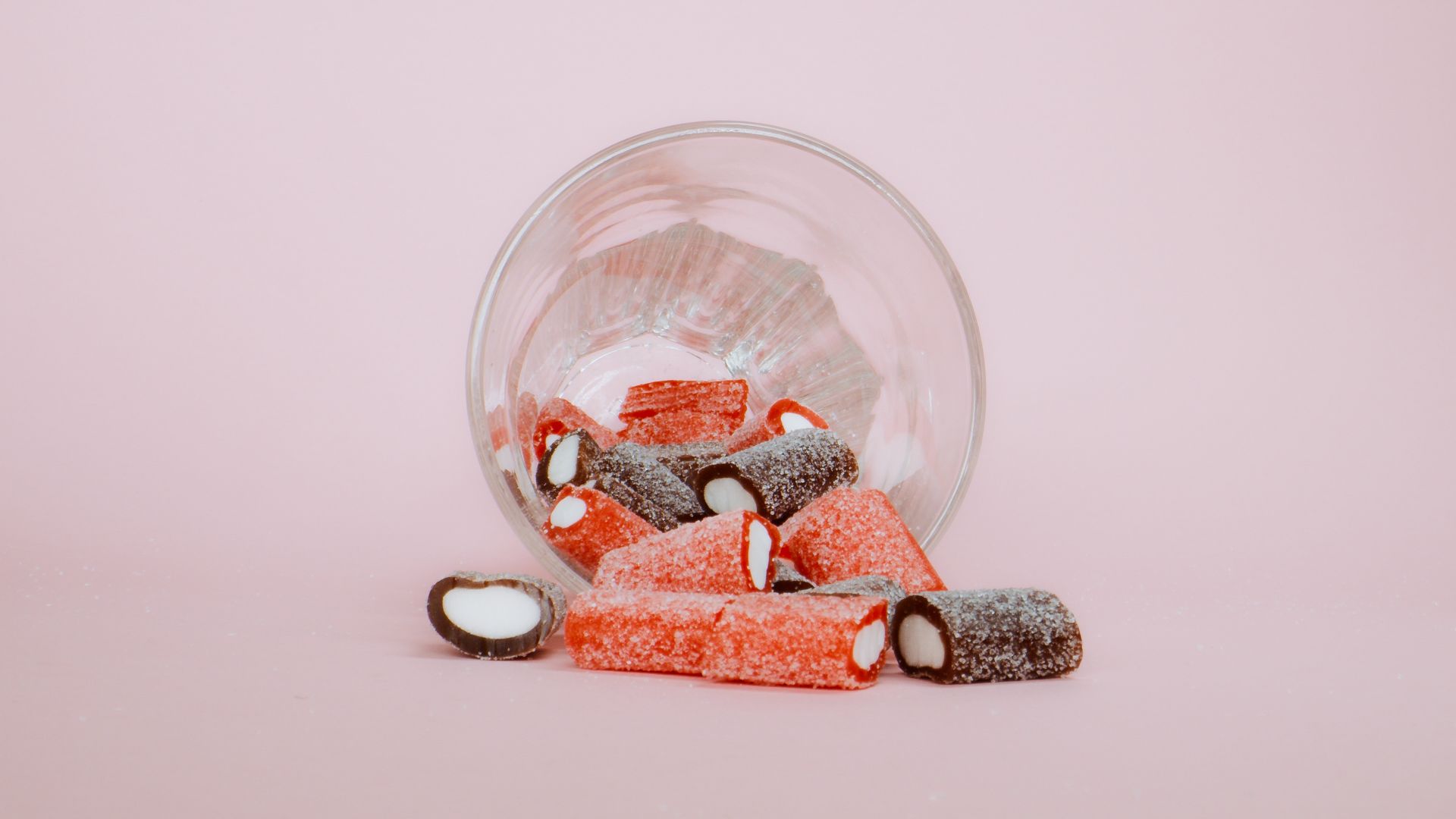 Vibrant, sugar-coated candies spill from a glass on a pastel pink surface.