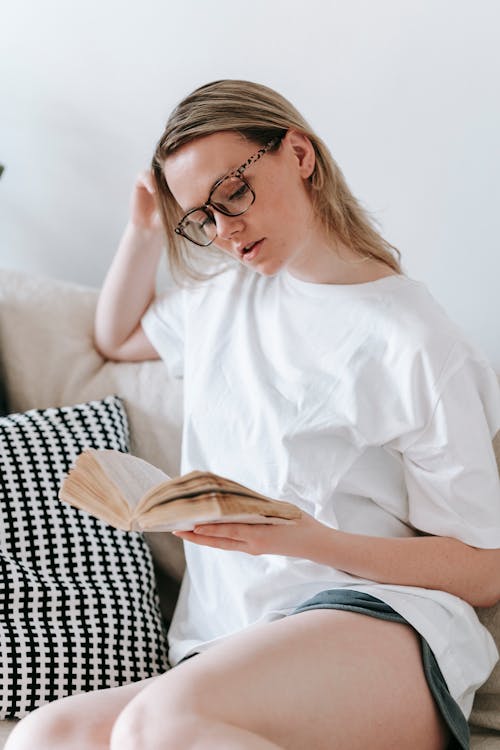 Serious woman in eyeglasses sitting on couch and reading book while leaning on hand