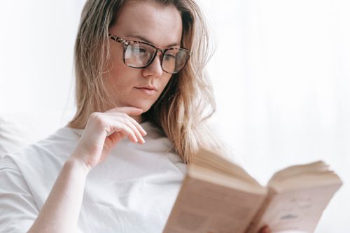 Free Low angle of crop concentrated female in eyeglasses touching chin while reading novel at free time Stock Photo