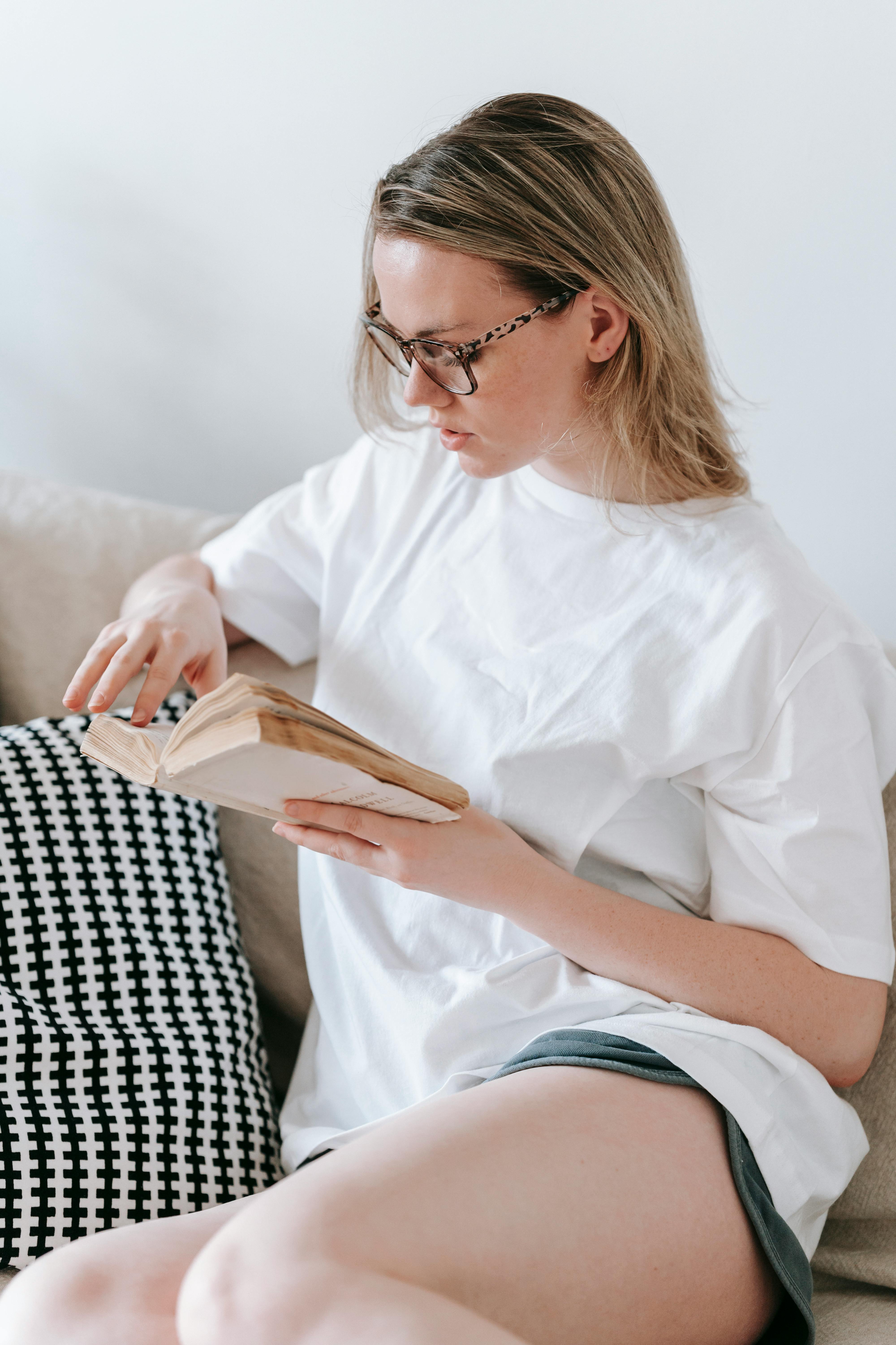 young woman in eyeglasses reading book on comfortable couch