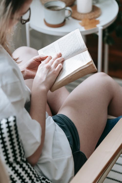 High angle of crop female in eyeglasses sitting in armchair and reading book attentively while relaxing at home