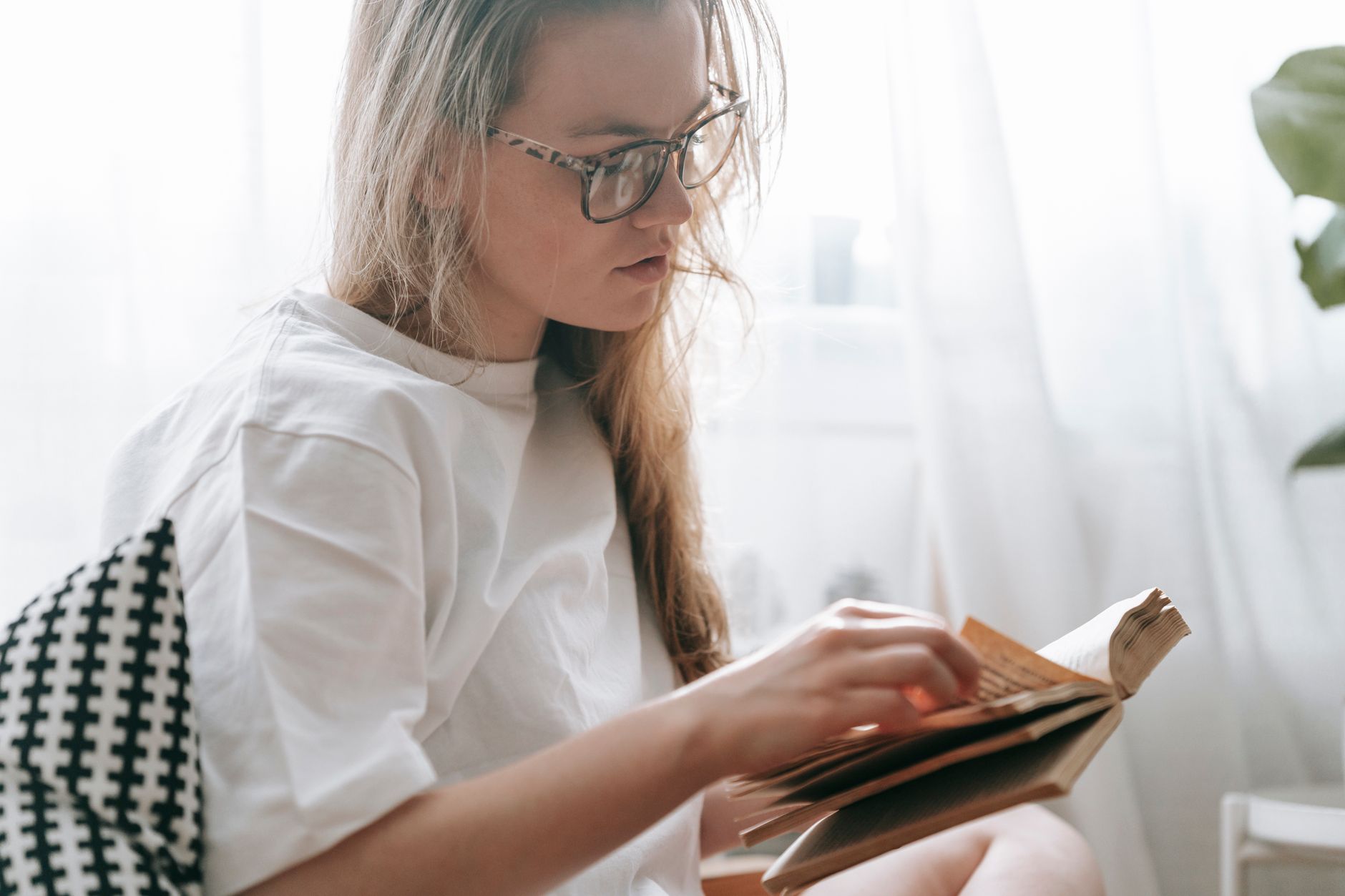 Side view of serious crop female with long hair in eyeglasses turning page of book while sitting near window