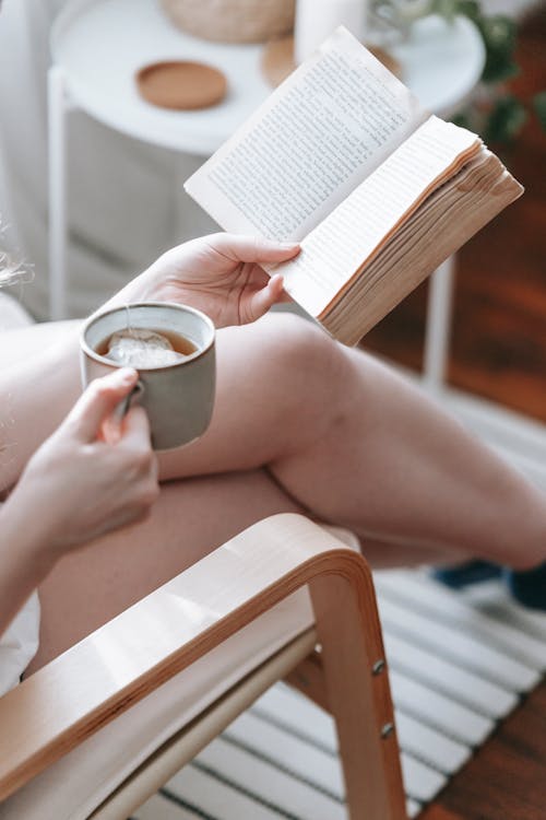 Free From above of crop anonymous female sitting with crossed legs and drinking tea while reading book at weekend Stock Photo