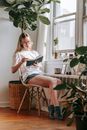 Young female in eyeglasses reading textbook while sitting on chair between tropical plants in house room