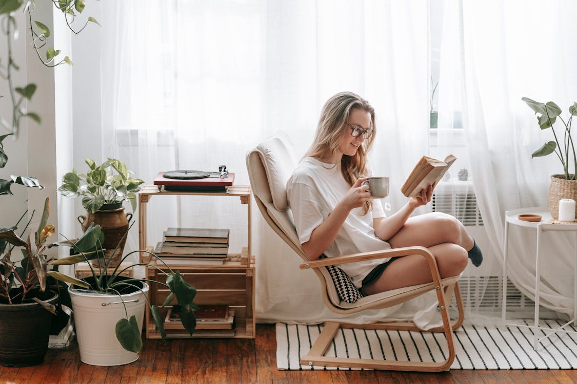 Cheerful young female in eyeglasses with cup of beverage reading textbook in armchair between potted plants in house room