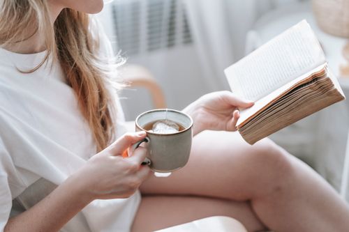 Free Crop anonymous female with crossed legs and cup of hot drink reading aged textbook in armchair at home Stock Photo