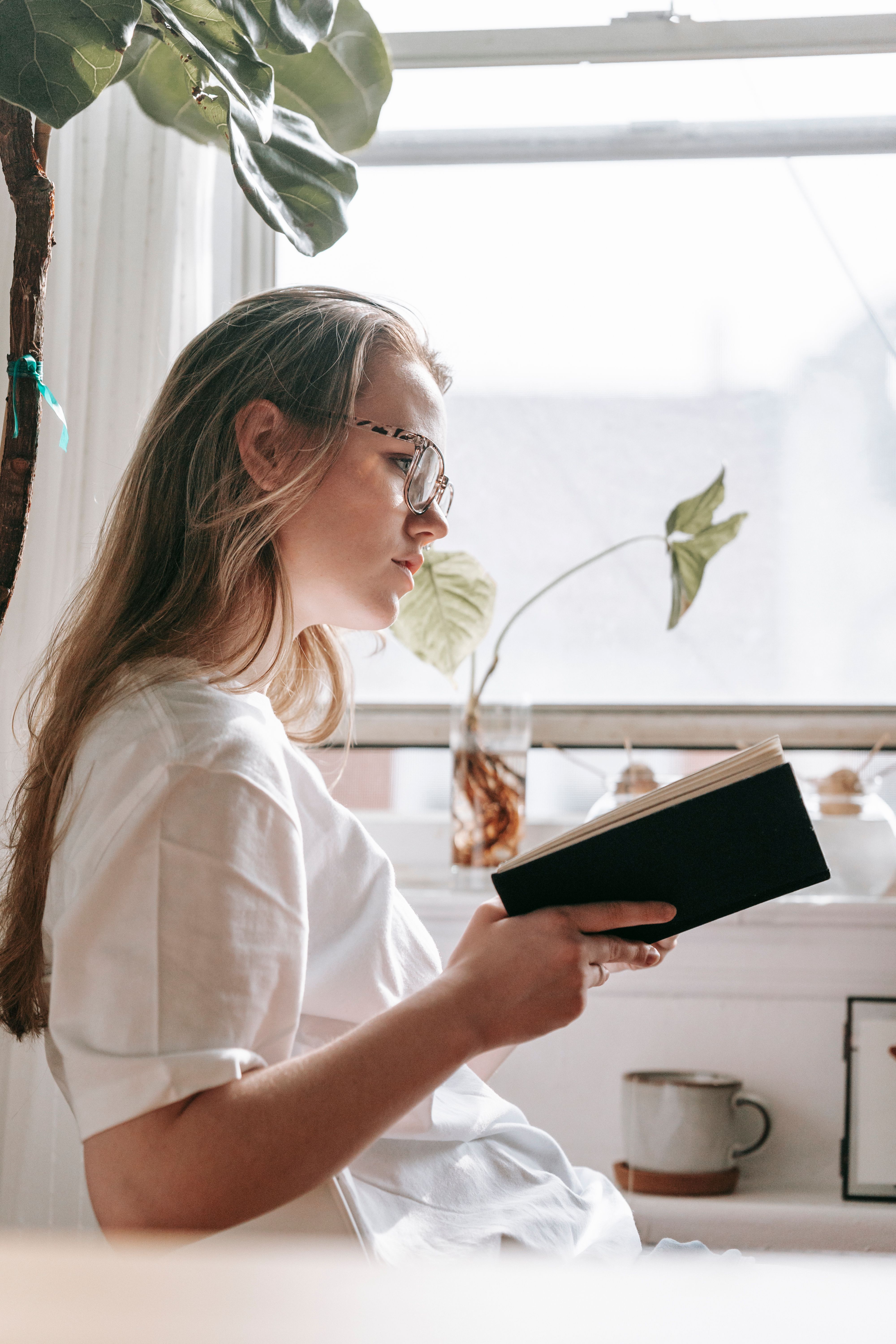A young woman in glasses reading beside a sunlit window with indoor plants