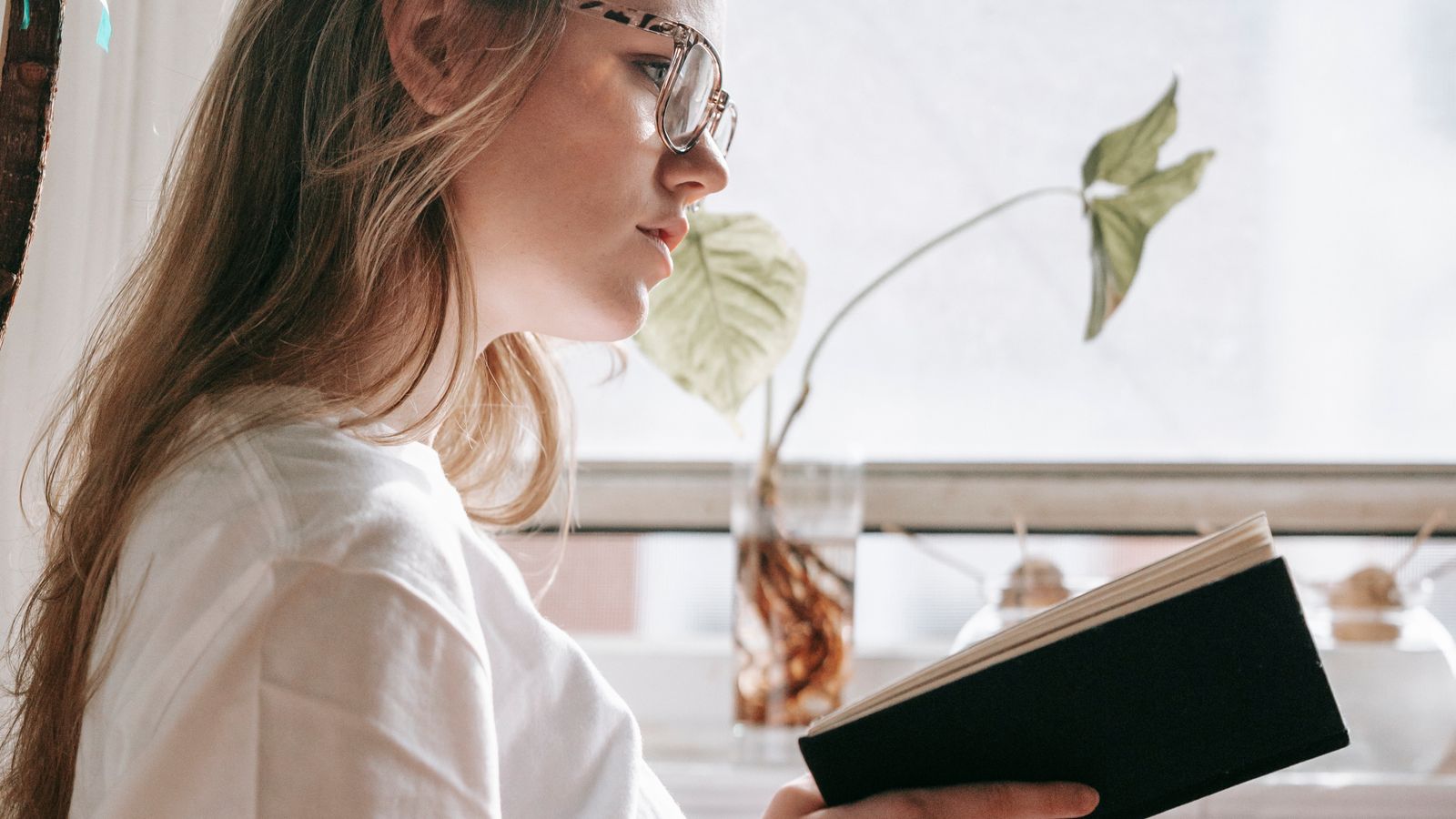 A young woman in glasses reading beside a sunlit window with indoor plants