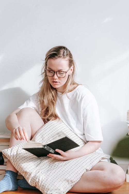 Free Young focused female in eyewear with cushion reading textbook while studying in house on sunny day Stock Photo