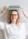 Sincere young female in eyewear and pile of textbooks on head looking away on sunny day on light background