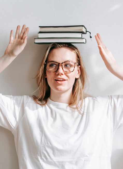 Sincere young female in eyewear and pile of textbooks on head looking away on sunny day on light background