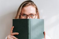 Clever young blond female in eyeglasses covering face with book while looking at camera on light background