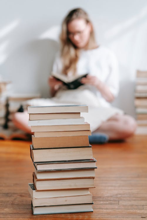 Heap of textbooks on parquet against woman preparing for exam in house on sunny day