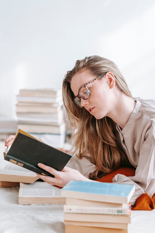 Side view of young focused woman in eyewear reading textbook while preparing for exam in house