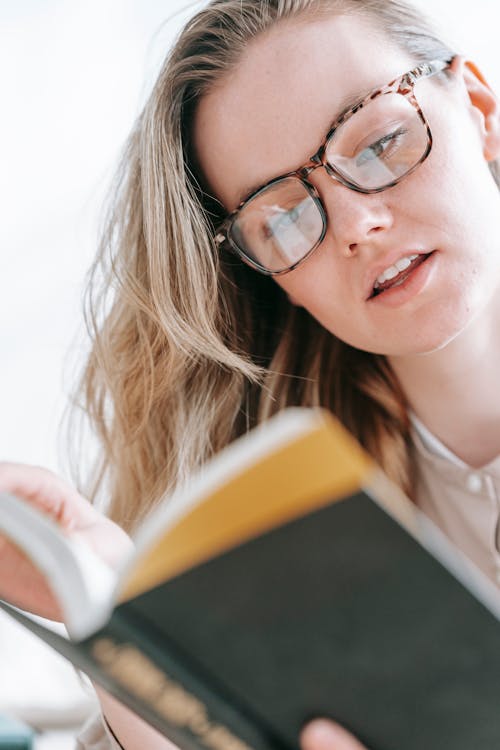 Low angle of crop concentrated female in eyeglasses reading interesting book attentively at free time