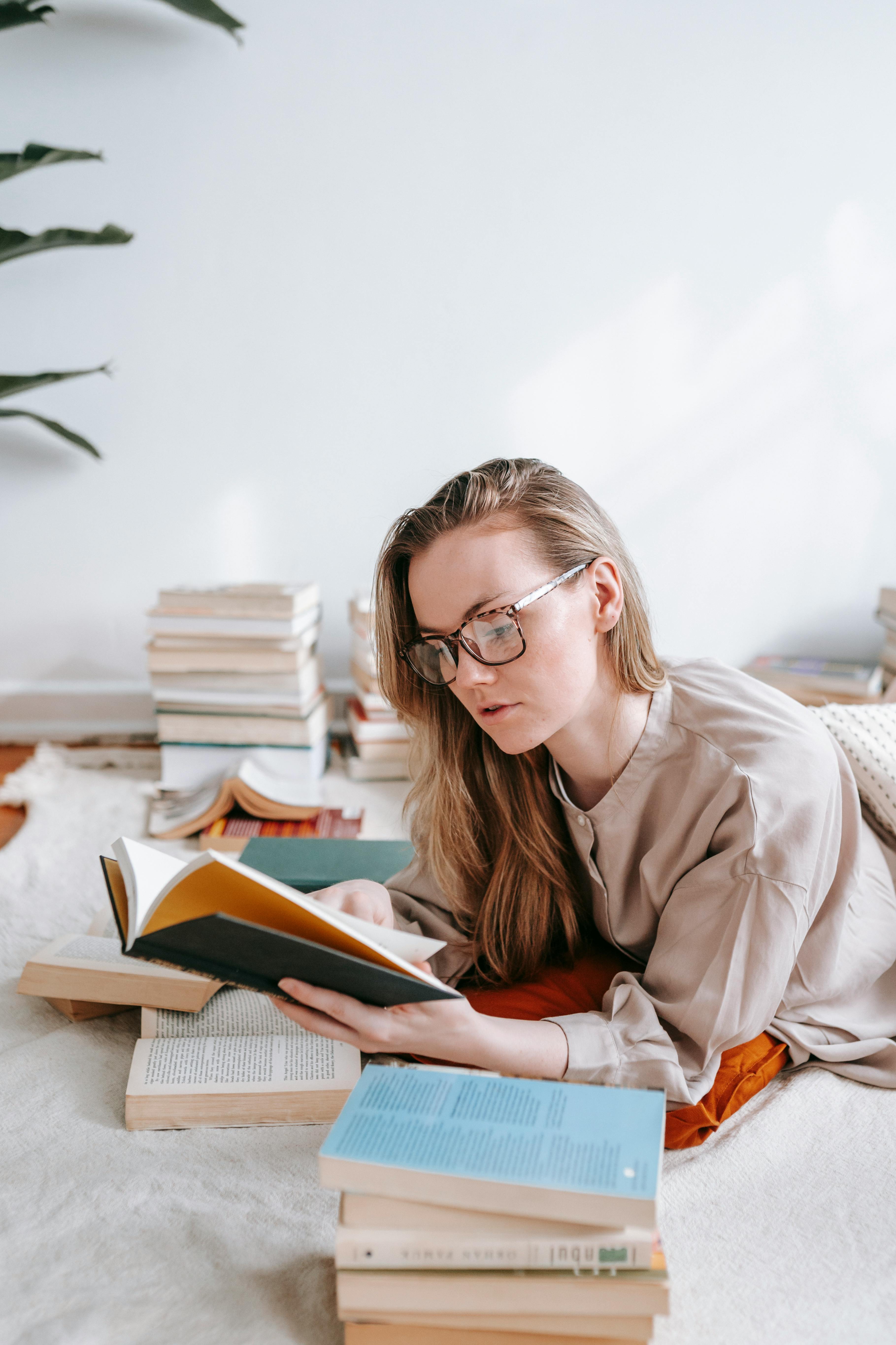 Free Woman with glasses reading in a cozy setting surrounded by books, embodying learning and leisure. Stock Photo