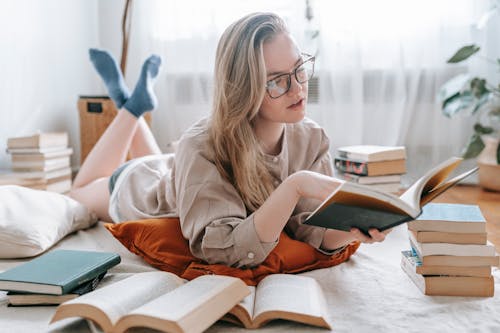 Full body of concentrated young female in eyeglasses reading book while preparing for studies at home