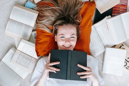 Free Top view of young female lying on floor among opened textbooks and books and smiling at camera Stock Photo