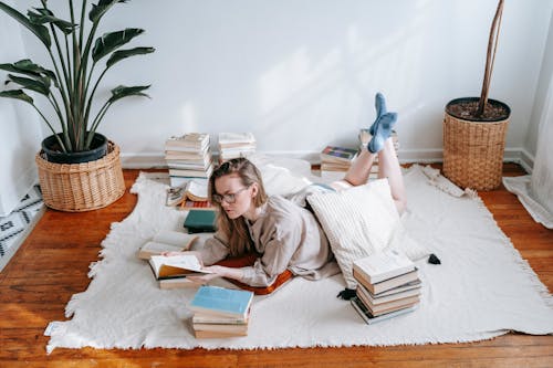 Full body of female in eyeglasses reading book while lying on plaid near heap of literature and green plants