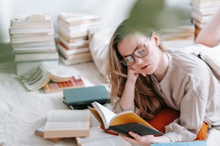 Concentrated young female in eyeglasses reading book while lying on white blanket near heap of literature in light room at home