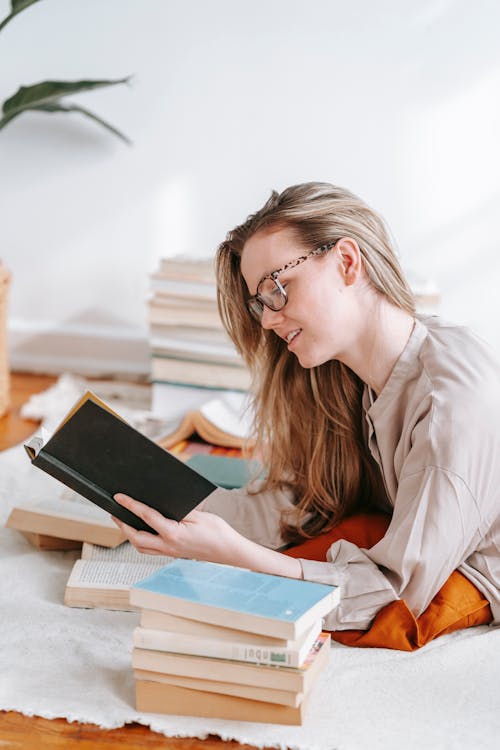 Side view of content female in eyeglasses reading book while lying on white plaid in floor with heap of literature