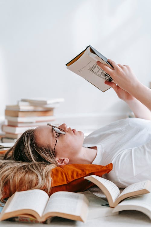 Side view of pensive young female in eyeglasses reading novel while lying amidst heap of literature in room during free time