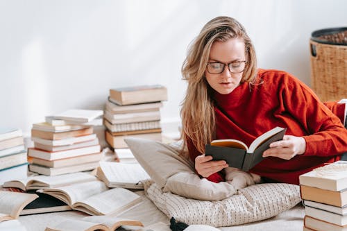 Attractive female in red sweater reading book while lying on floor with pillows and heap of literature in light room