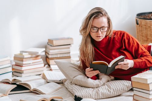 Free Attractive female in red sweater reading book while lying on floor with pillows and heap of literature in light room Stock Photo