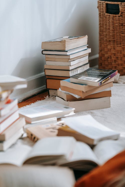 Collection of books placed on white soft textile on floor near wall in light room with wicker basket at home