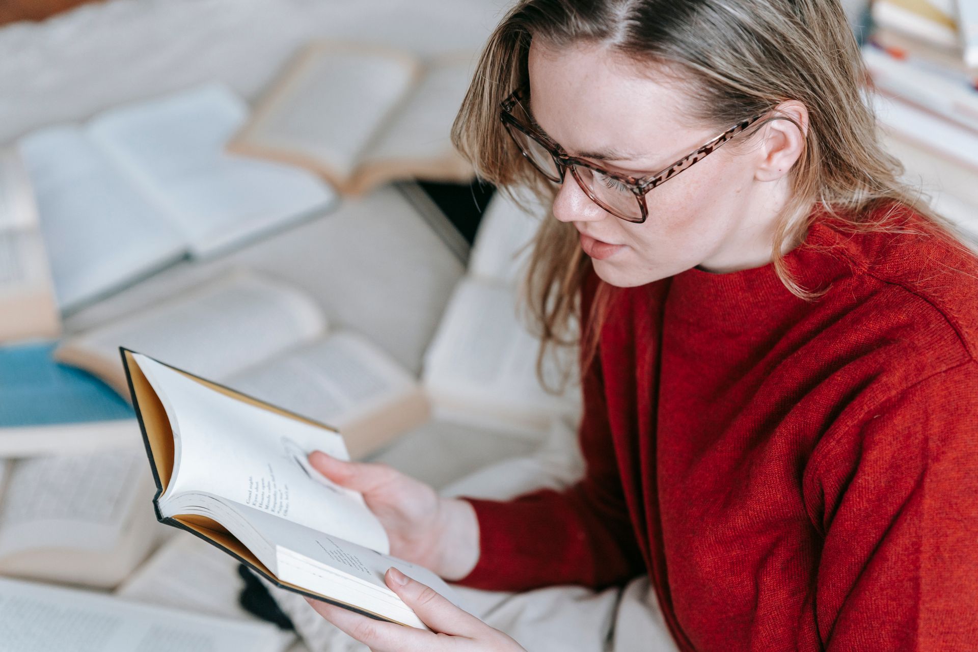 From above side view of crop concentrated female in red outfit reading book while sitting near heap of literature in light room
