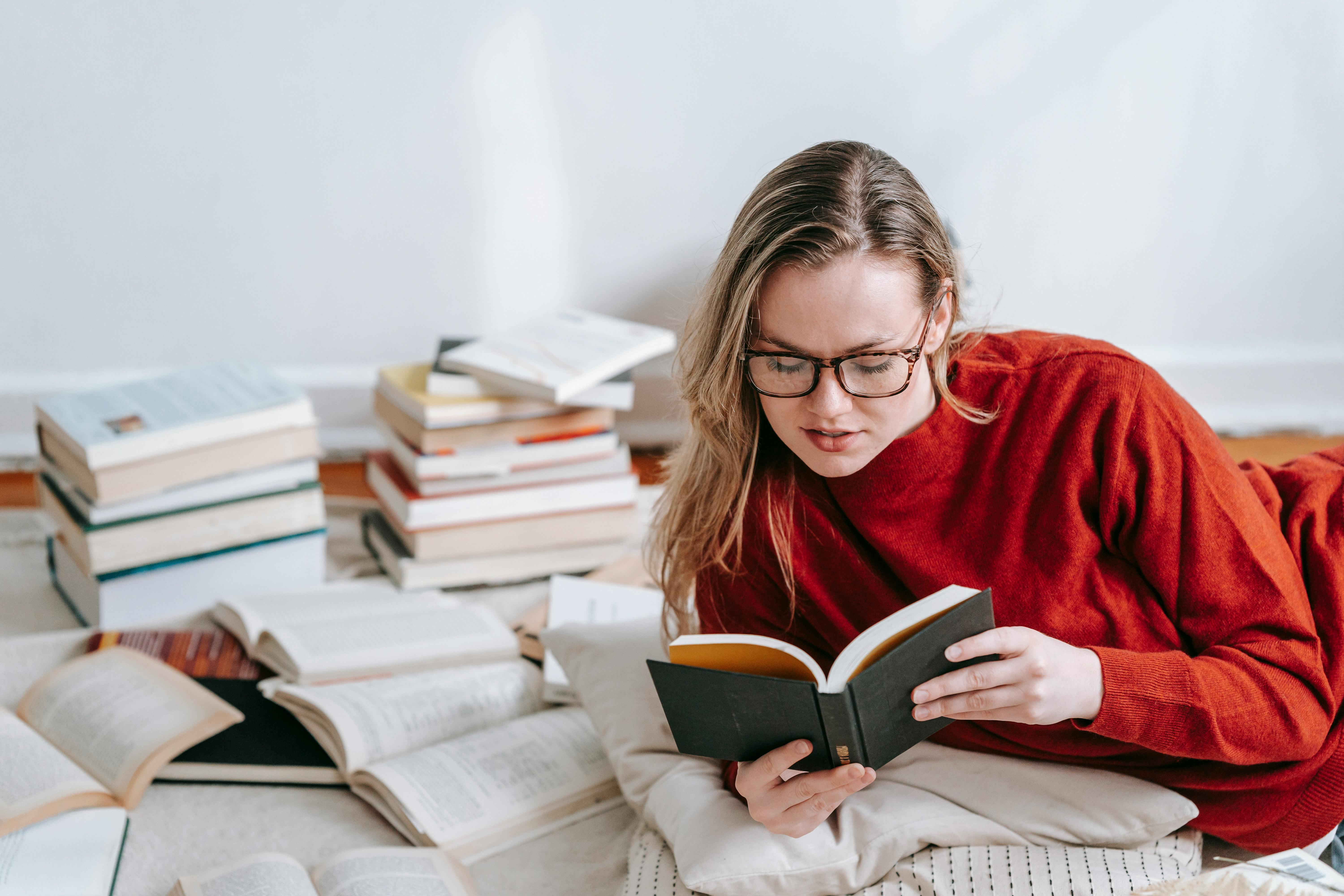 Free A woman in a red sweater reading a book on the floor with stacks of books around her indoors. Stock Photo