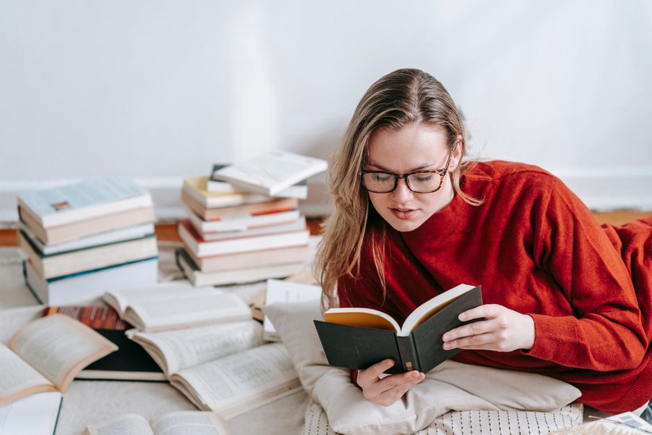 Intelligent female in eyeglasses reading book while lying on on floor with cushion and pile of literature in light room
