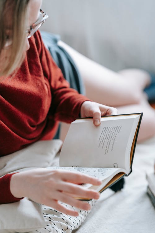 Crop unrecognizable female in eyeglasses reading book while lying on white blanket on floor in light room during free time on blurred background