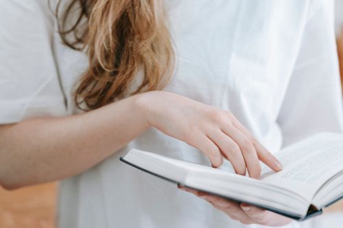 Free Crop anonymous female in white t shirt with long hair reading book in hand while spending free time in light room Stock Photo