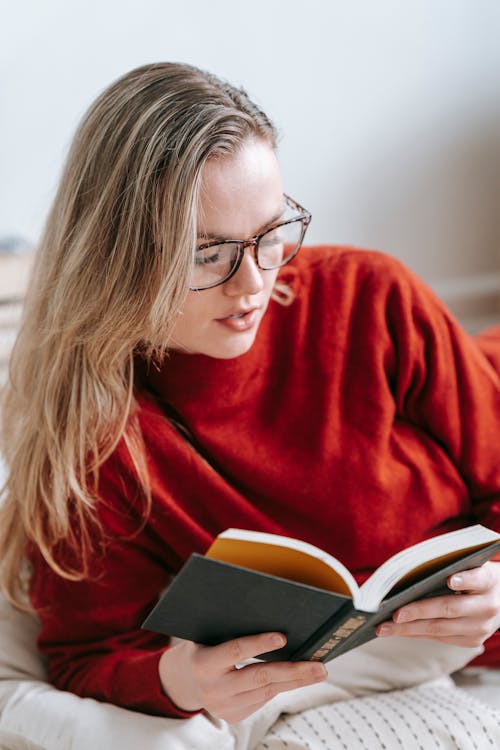 Free Concentrated intelligent female in eyeglasses reading interesting book while lying on floor with pillows in light room during free time Stock Photo