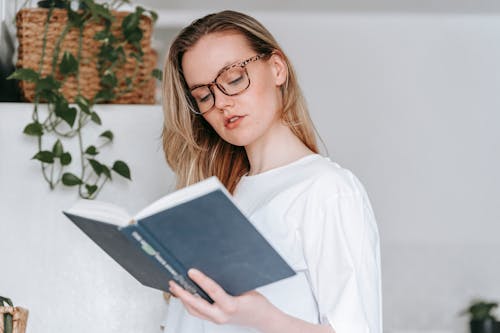 Concentrated young female in eyeglasses and white garment reading book while standing in light kitchen with fridge and green plant