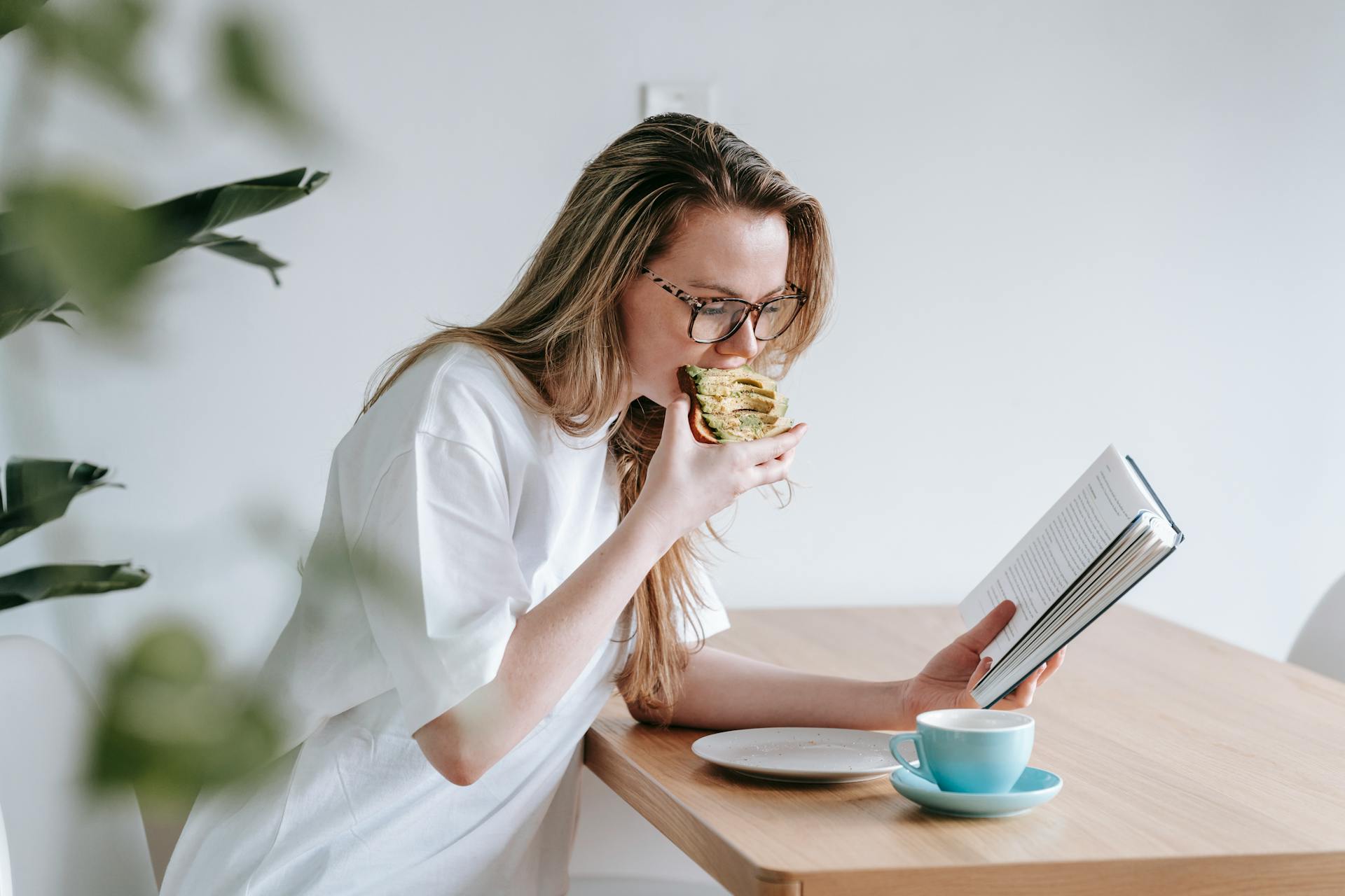 Side view of smart female in eyeglasses with long hair studying book at table with cup and plate