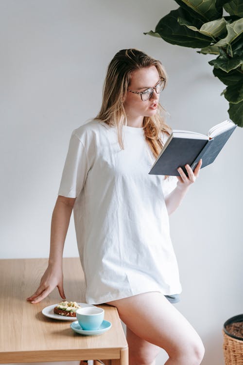 Free Attractive young woman in white t shirt sitting at table and reading interesting book while having coffee and toast Stock Photo