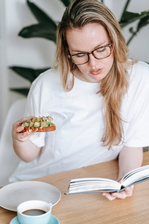 Free Young female in white t shirt sitting at table with cup of coffee and reading interesting book while having avocado toast for breakfast Stock Photo